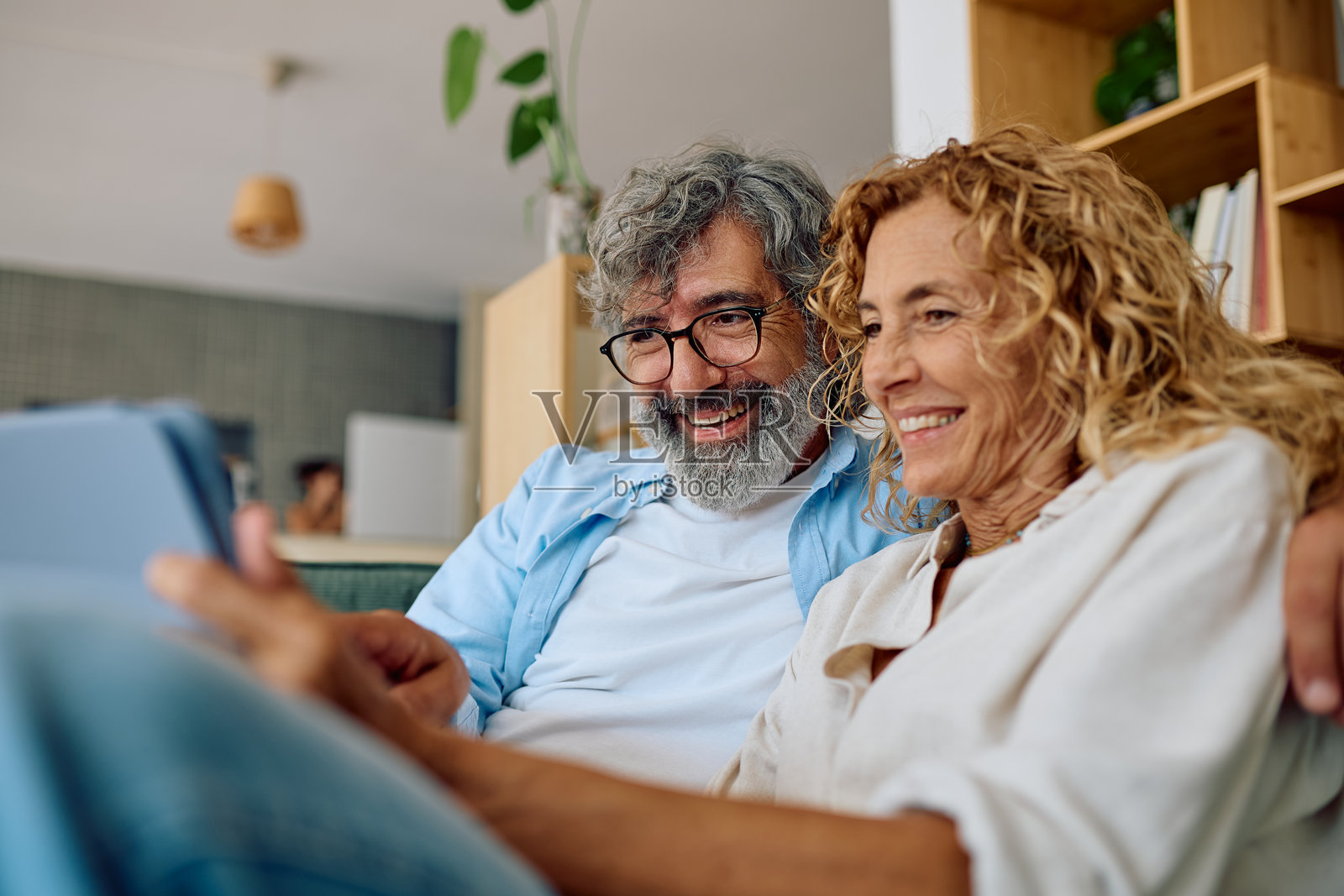 快乐的老年夫妇在家中使用平板电脑，放松地坐在沙发上并微笑。照片摄影图片