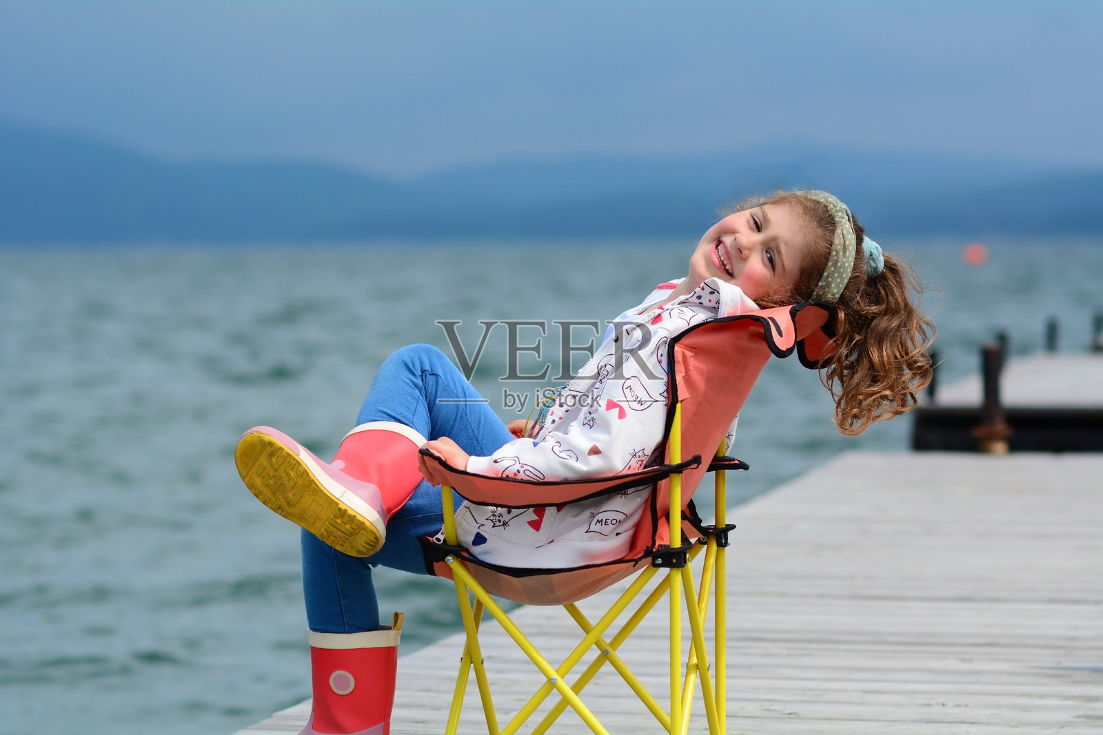 小女孩坐在椅子上，背景是湖景的木码头。照片摄影图片