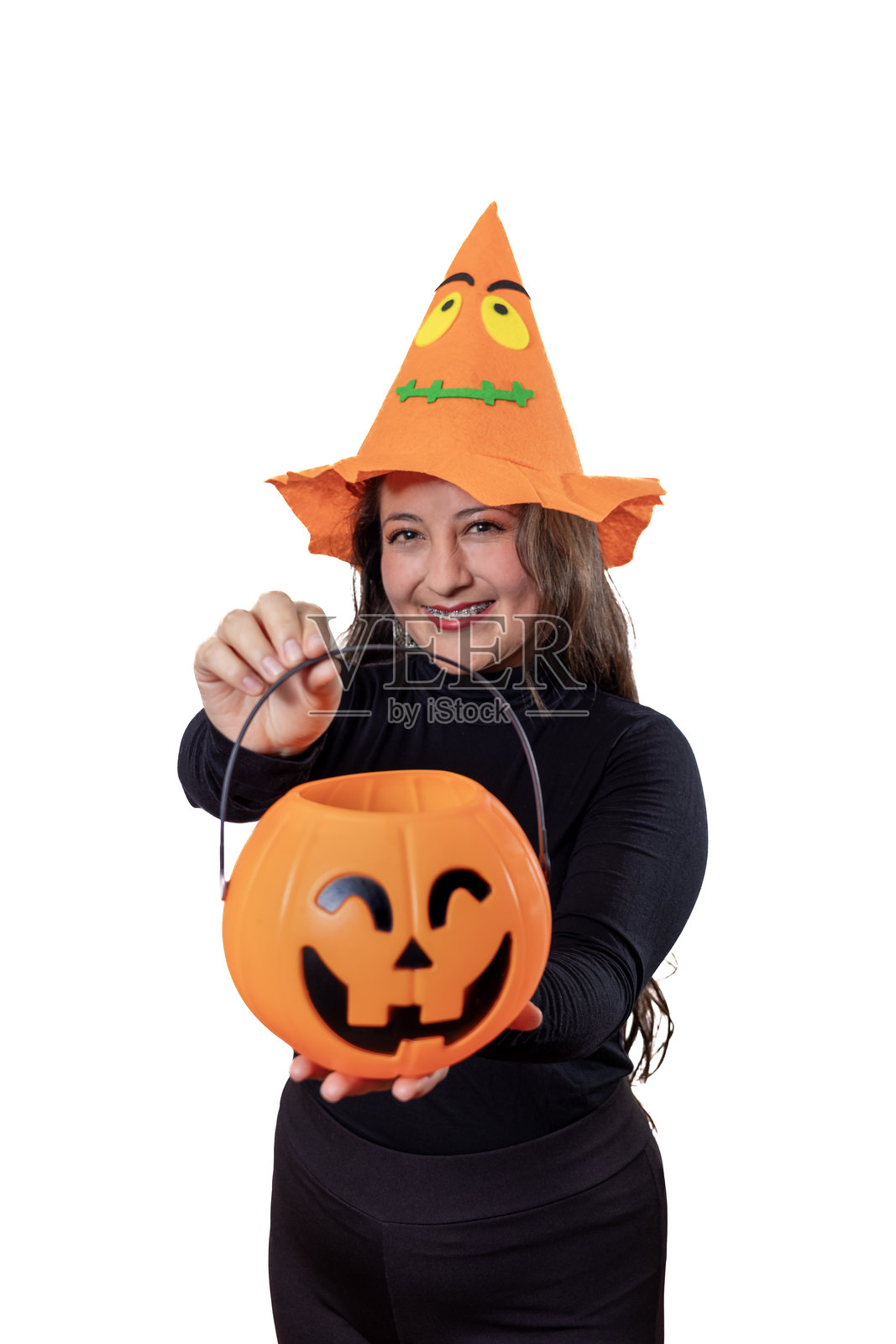 年轻女子戴着南瓜帽，穿着黑色衬衫，对着镜头微笑，手里拿着橙色南瓜灯糖果桶，庆祝万圣节。照片摄影图片