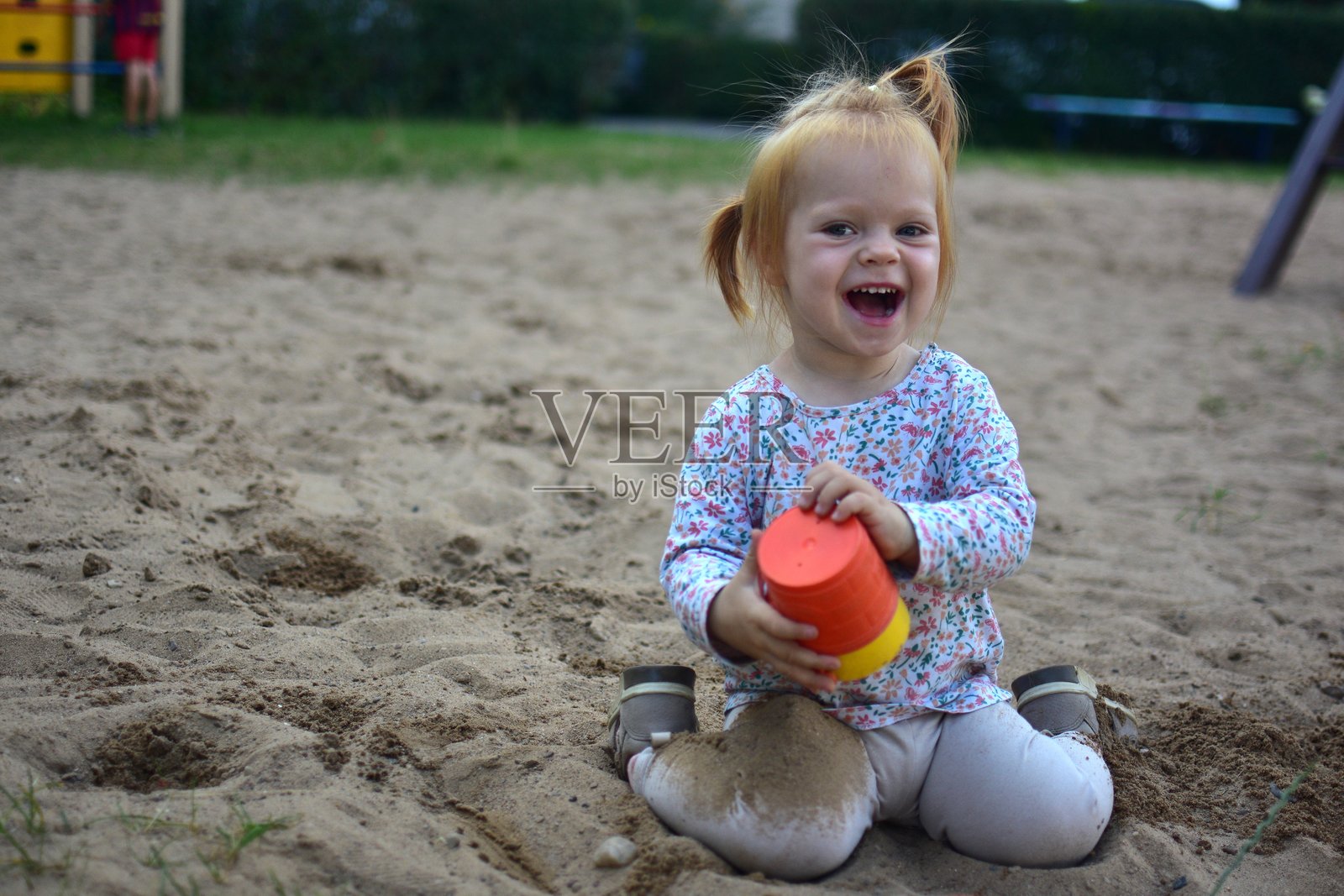 快乐的幼儿女孩在沙坑里大笑，手里拿着色彩缤纷的水桶，享受户外玩耍的乐趣，在愉快的童年时光里散播着欢乐与纯真。照片摄影图片