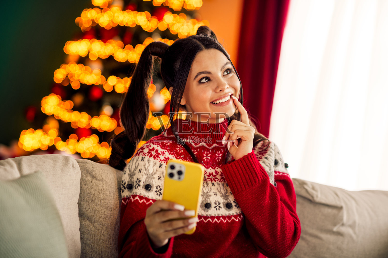 一位年轻女子在室内圣诞树旁，身着节日舒适毛衣，手持智能手机，微笑着享受假日的时光。照片摄影图片