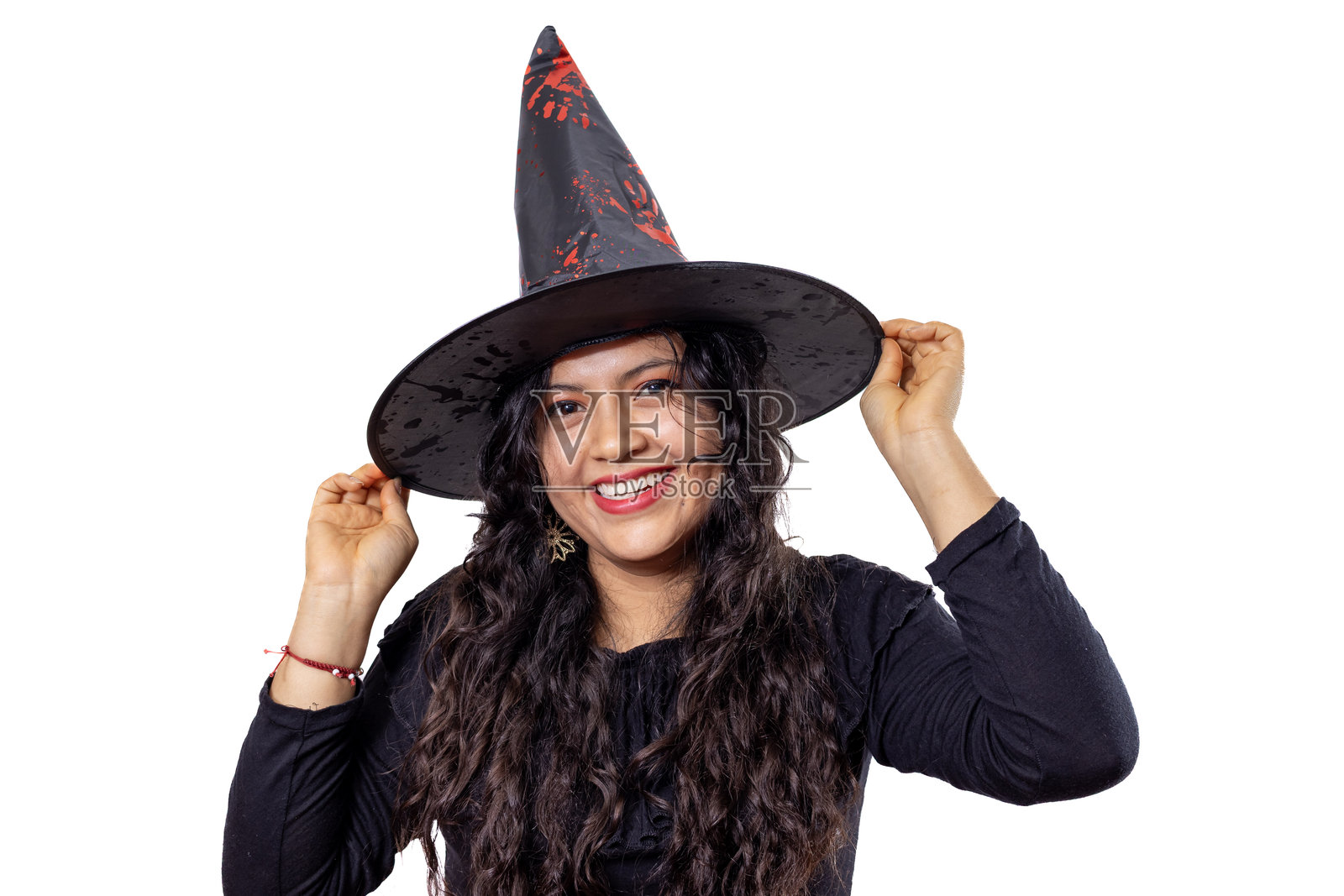 年轻的拉丁裔女性，身着万圣节服装，在白色背景前，脸上带着笑容，手中拿着一顶沾有假血的女巫帽的帽檐，庆祝这个诡异的节日。照片摄影图片