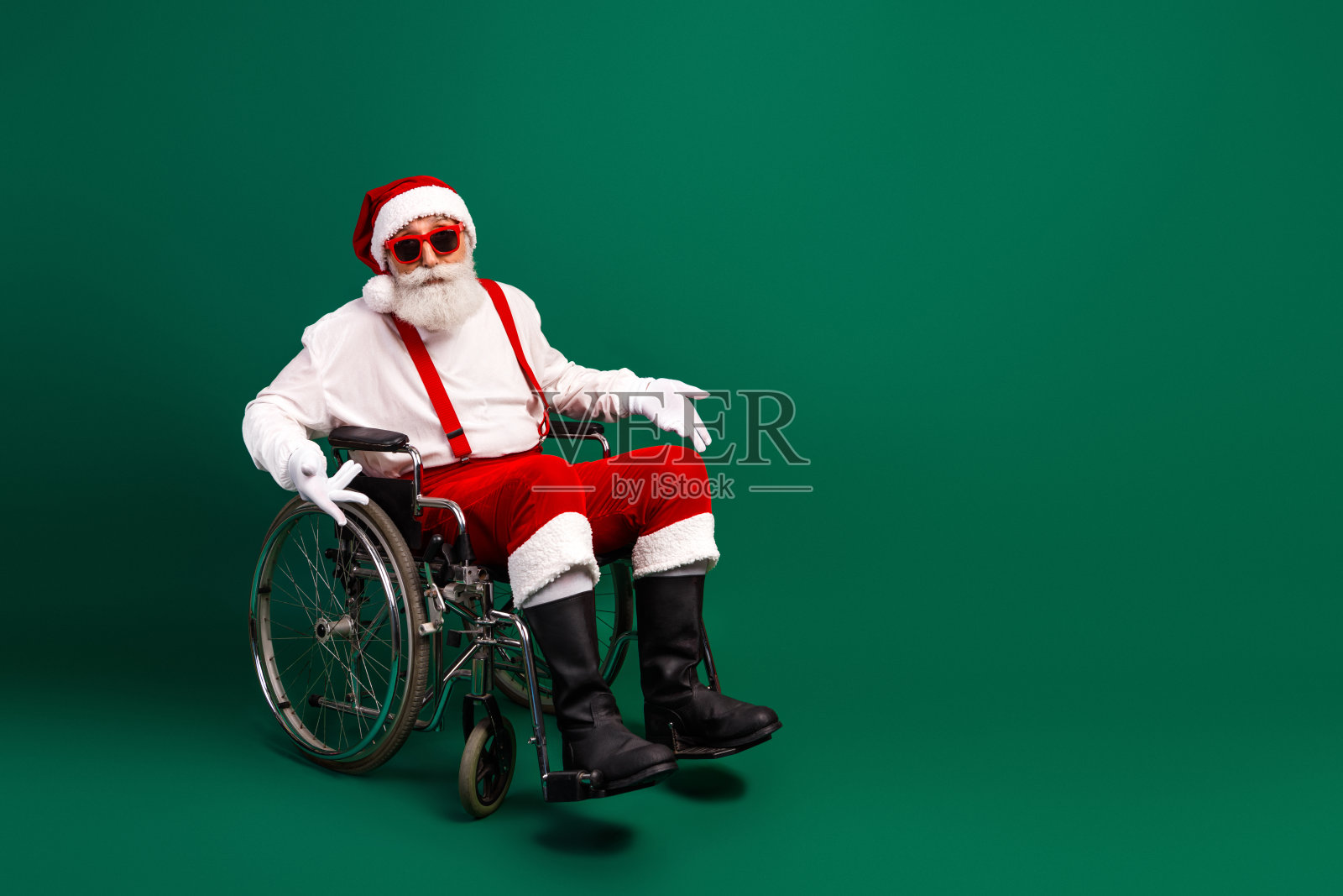 坐着轮椅的圣诞老人，戴着太阳镜，穿着红色服装，在绿色背景上散发出欢乐喜庆的氛围照片摄影图片