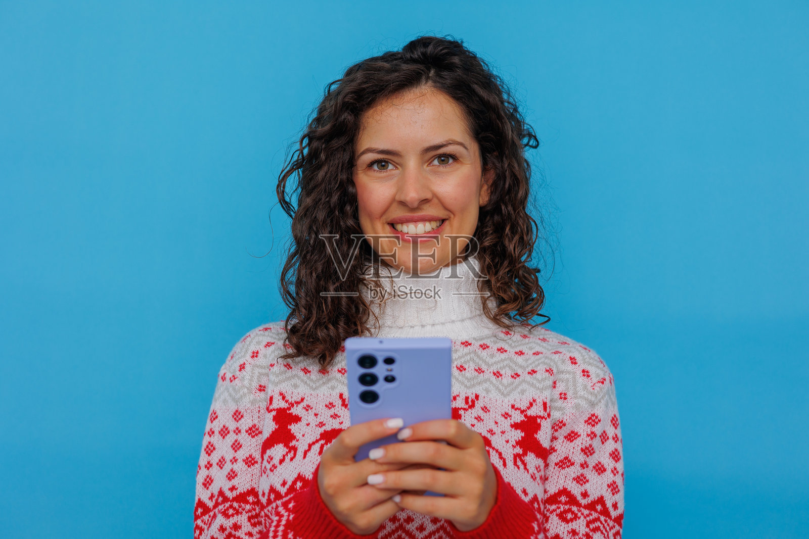 一位留着卷发的快乐女子，穿着舒适的毛衣，微笑着用智能手机发短信，充满节日冬日氛围和在线连接感。照片摄影图片