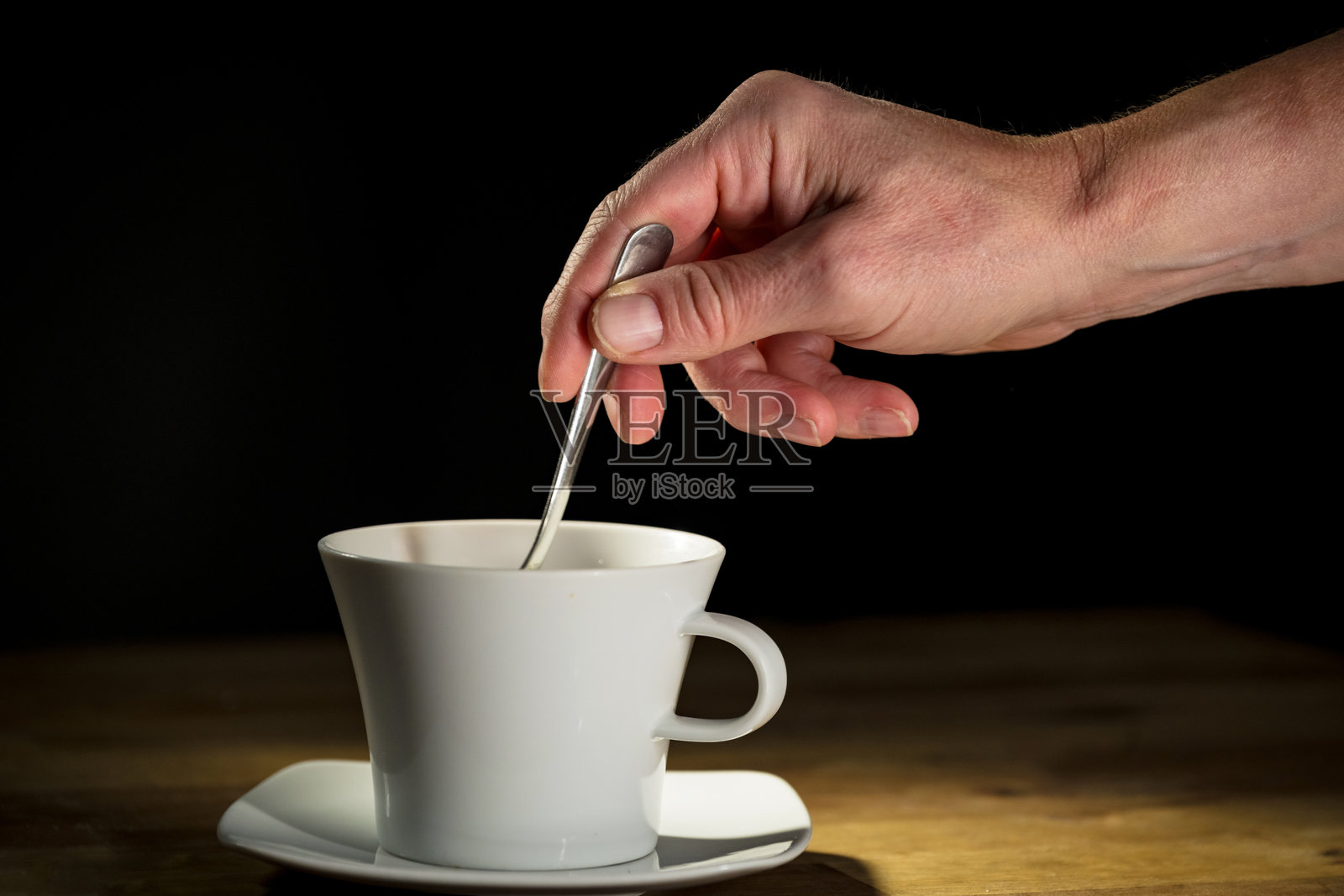 男士手持茶匙向白色杯子中加糖，背景为黑色。照片摄影图片