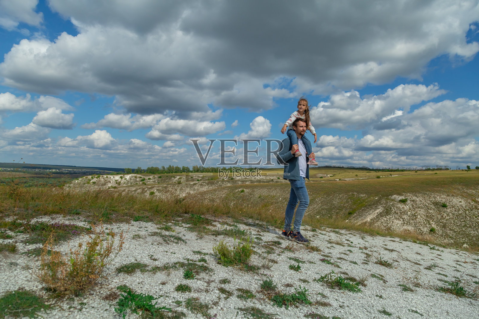 小女孩坐在父亲的肩膀上，手牵着手，望着远方，享受着大自然。父亲带着儿子在户外散步。照片摄影图片