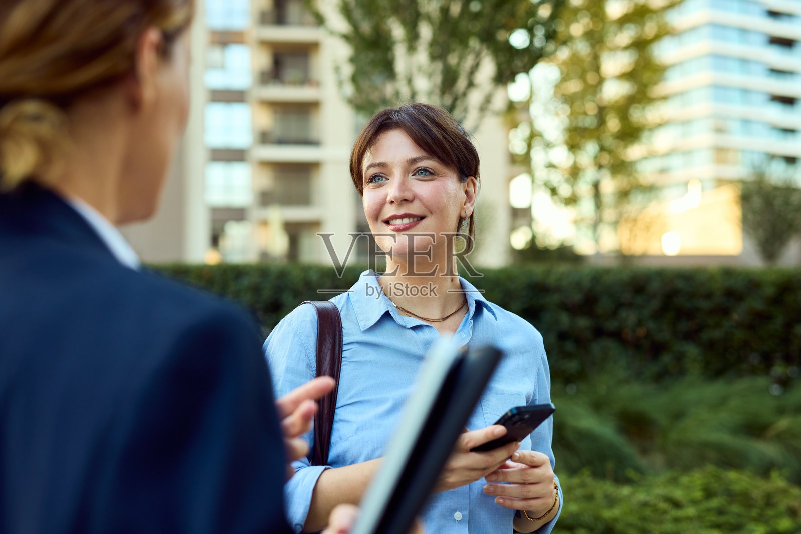 自信的女性在商务社交活动中，一边使用手机和文件，一边进行户外商务对话。照片摄影图片