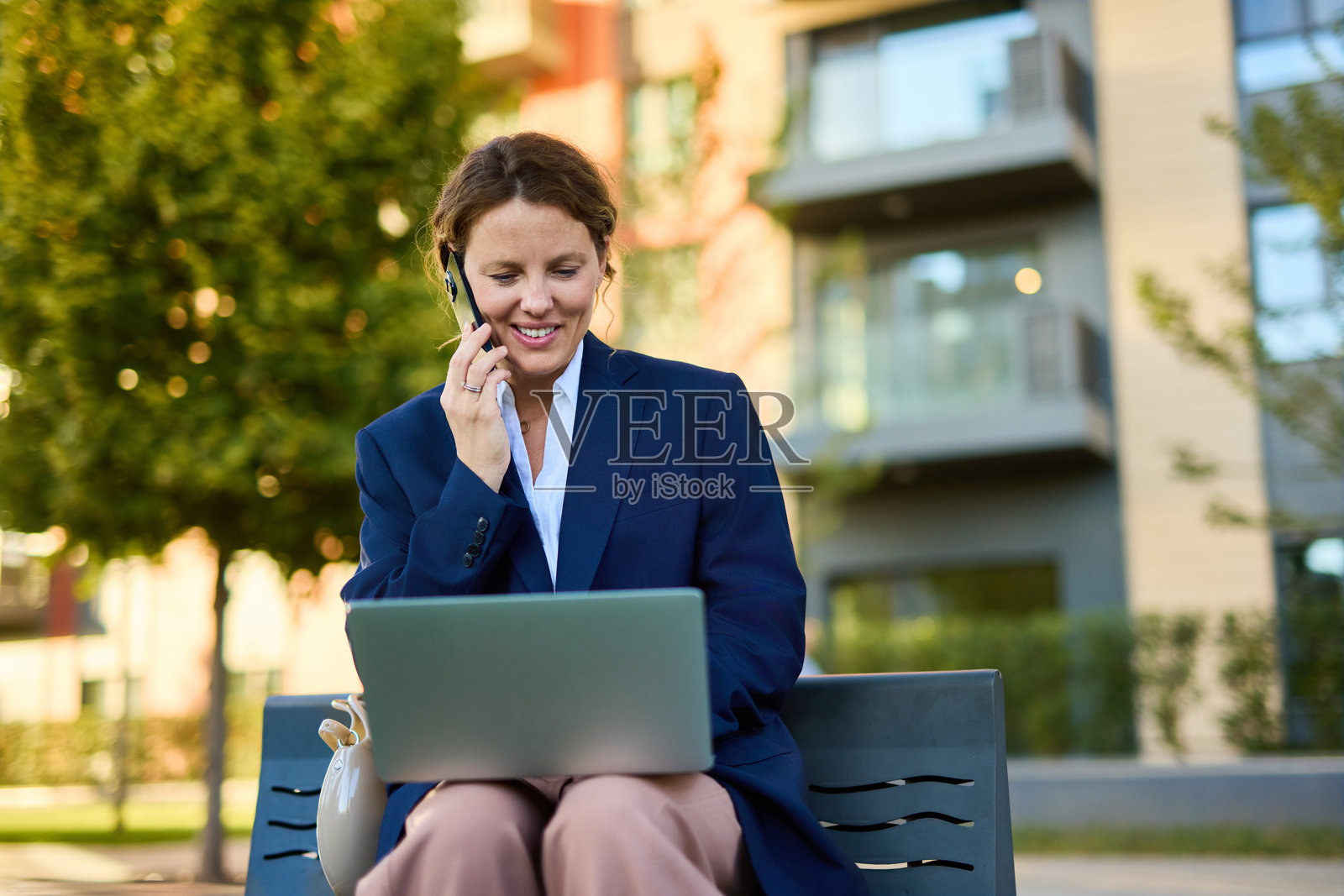 都市公园里，女商人一边打电话一边用笔记本电脑工作，展现了移动办公的场景。照片摄影图片