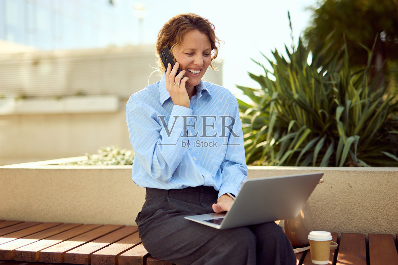 公园长椅上，一位快乐的职业女性在户外使用手机和笔记本电脑。照片摄影图片