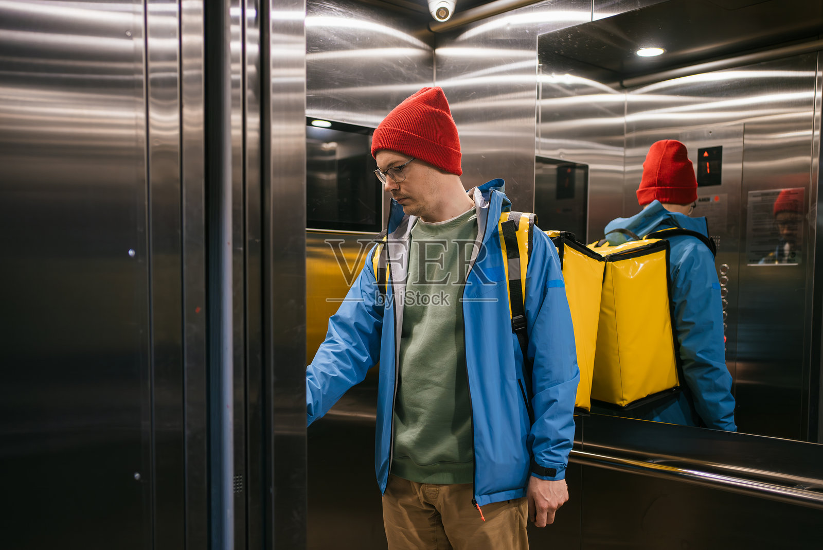 穿着红色羊毛帽和蓝色夹克的送餐员正在按电梯按钮，他背着黄色的保温背包，提供送餐服务照片摄影图片