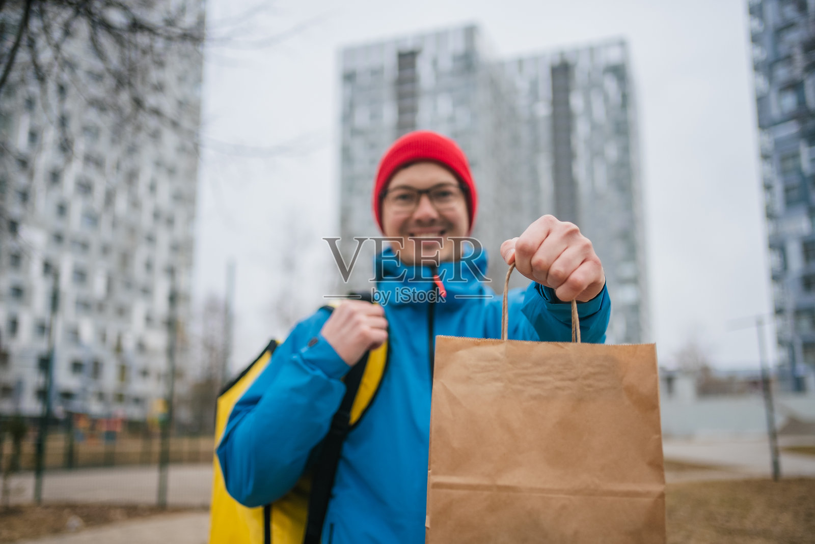 穿着蓝色夹克的送餐员背着保温背包，在有公寓楼的居民区运送纸袋餐食。照片摄影图片