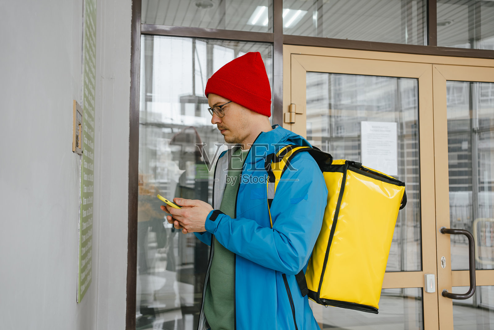 身穿蓝色夹克的送餐员使用智能手机，背着黄色保温背包，在建筑入口附近查看地址，同时配送食品。照片摄影图片