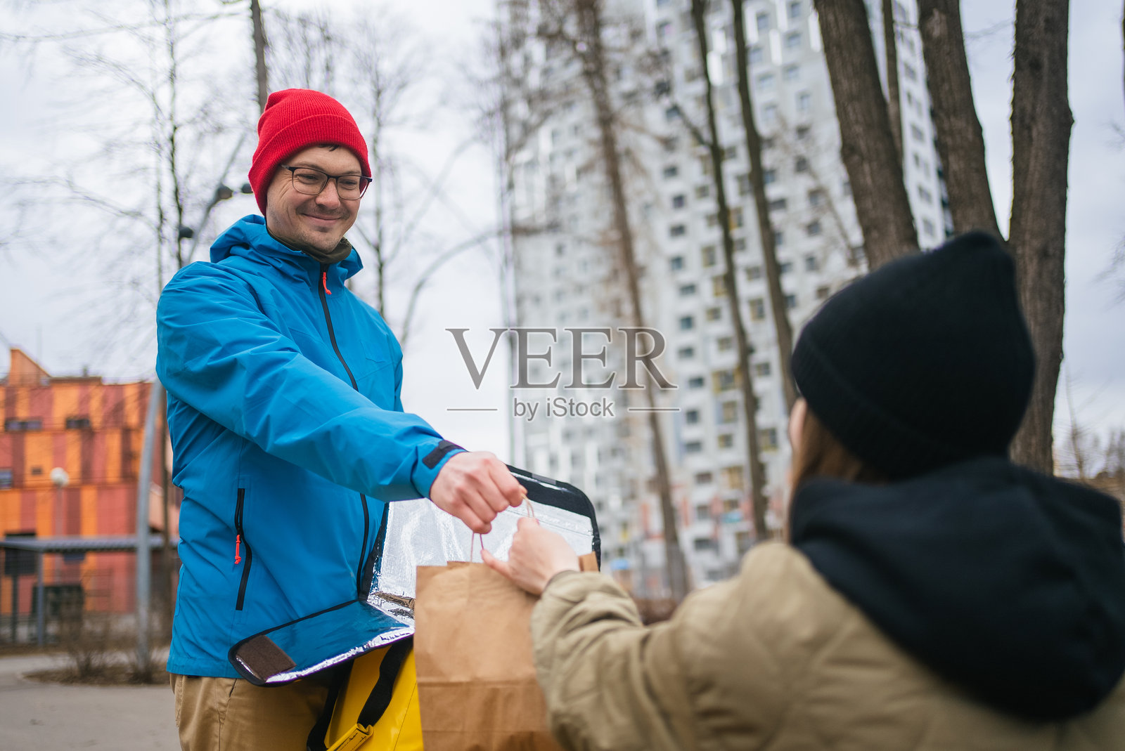 送餐员在繁忙的城市街道背景下，将装有外卖的纸袋递给顾客，背景中有现代建筑和绿树点缀的城市环境。照片摄影图片