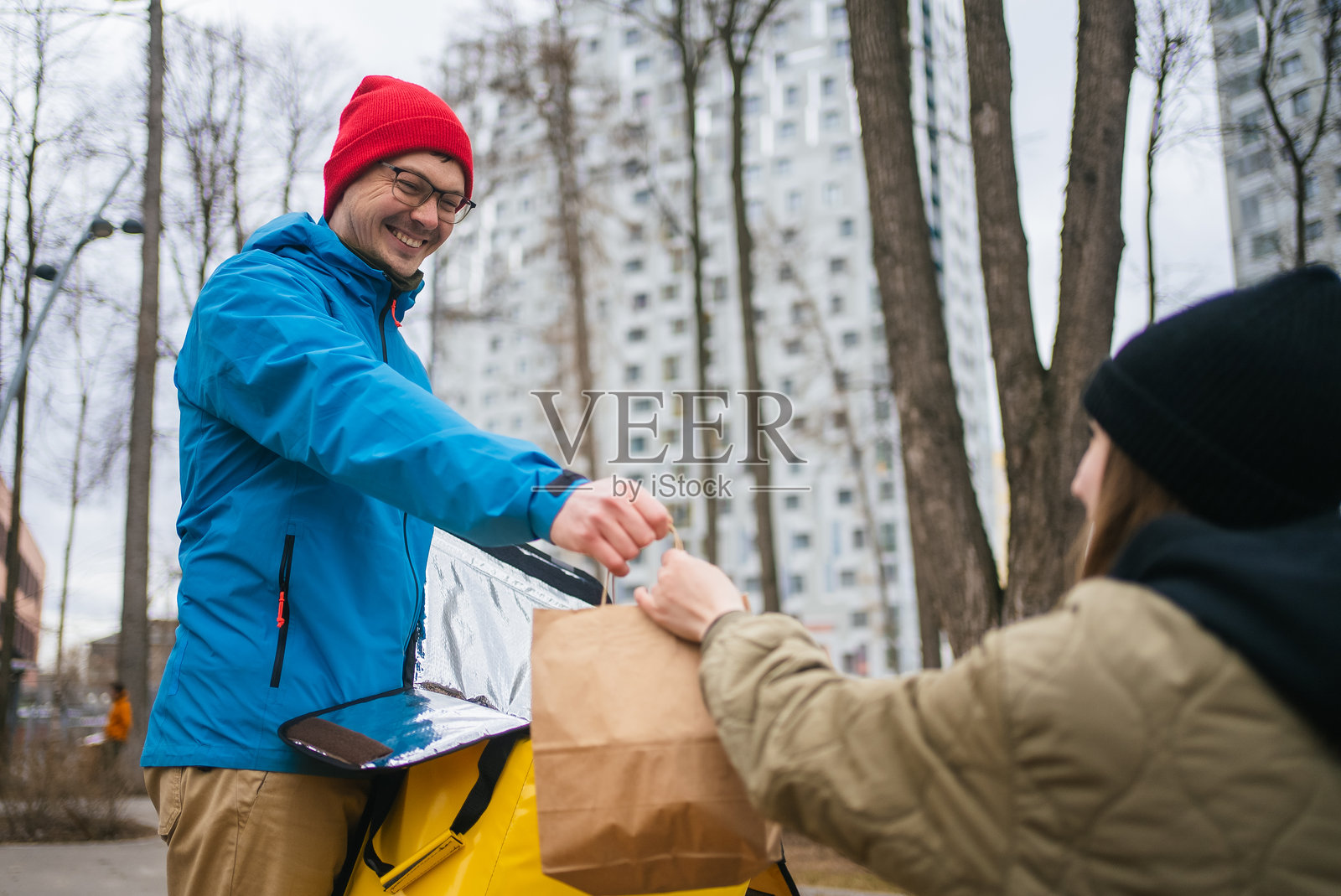 快乐的快递员将装有外卖的纸袋递给在城市绿化旁边的女性收件人，背景是繁华的城市景观。照片摄影图片