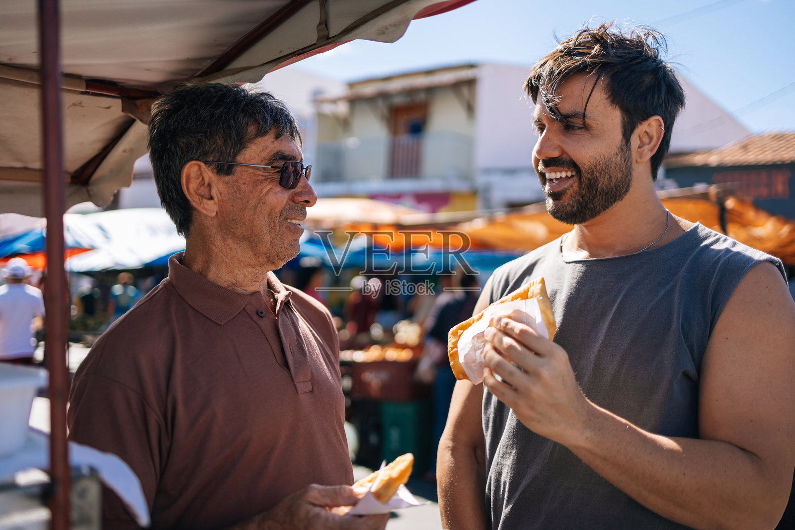老年父亲和成年儿子在露天市场吃着巴西传统油炸小吃 Pastel照片摄影图片