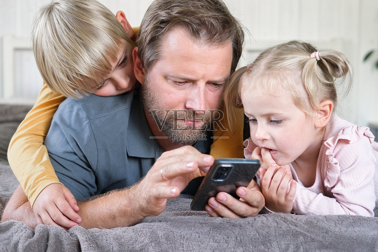 年轻的爸爸和孩子们一起使用智能手机。男人在看手机里的内容。快乐的父亲、女儿和儿子正在用手机玩耍，对着有趣的视频、度假照片、手机游戏微笑。照片摄影图片
