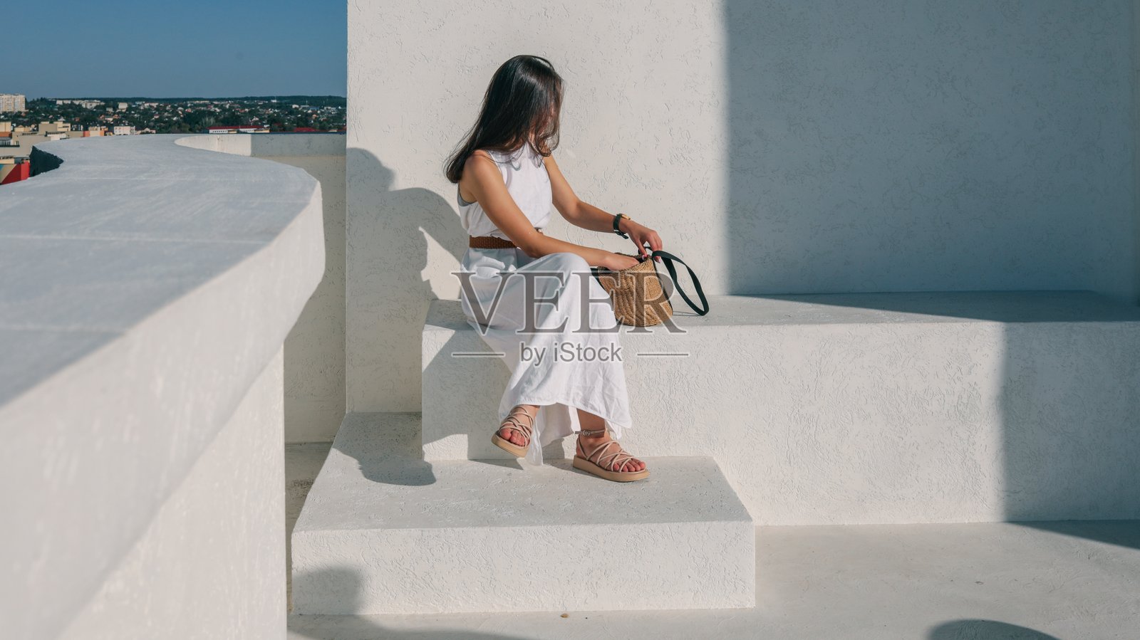 身穿白裙的女子坐在白色台阶上，旁边放着一个草编包照片摄影图片