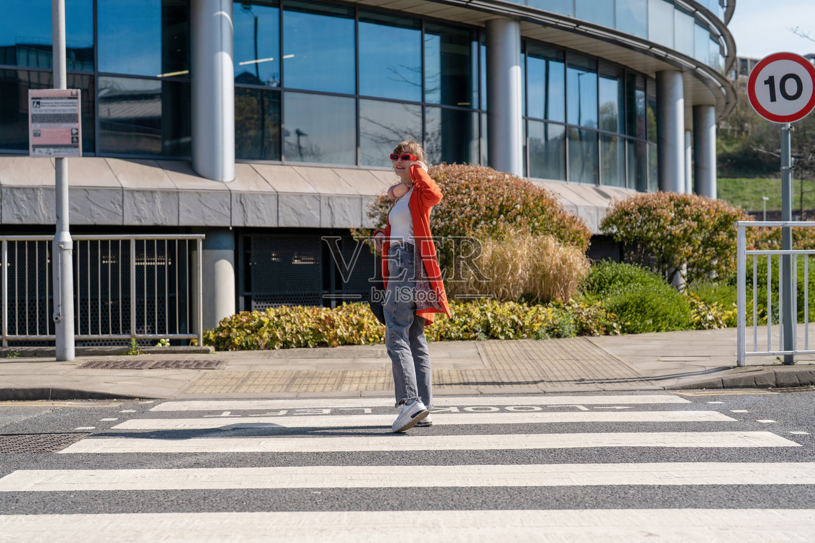 英格兰晴朗的一天，一位身穿红色开衫的年轻女子正在现代建筑前的斑马线上过马路。照片摄影图片
