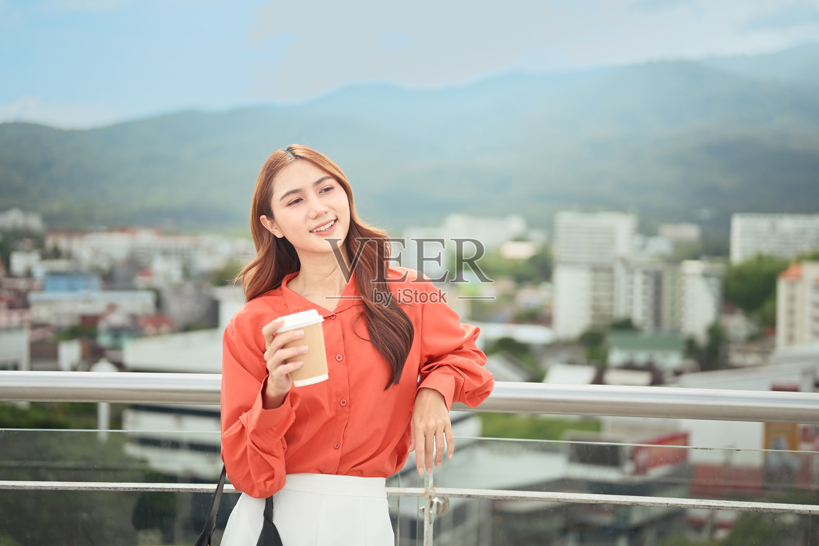 一位女士在屋顶露台上享受咖啡，背景是城市建筑和远山，她面带微笑。照片摄影图片