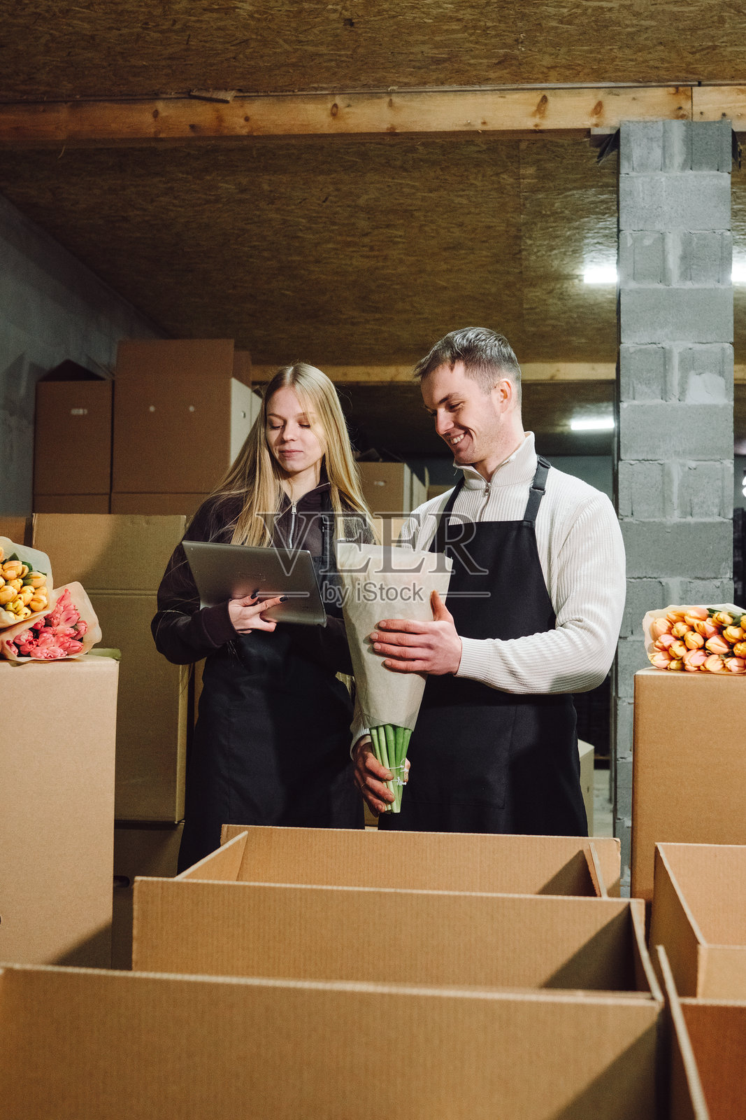 年轻女子和男子穿着黑色围裙，在堆满纸箱和鲜花的储藏区准备花艺作品，展现了花卉生意运营中的团队合作。照片摄影图片