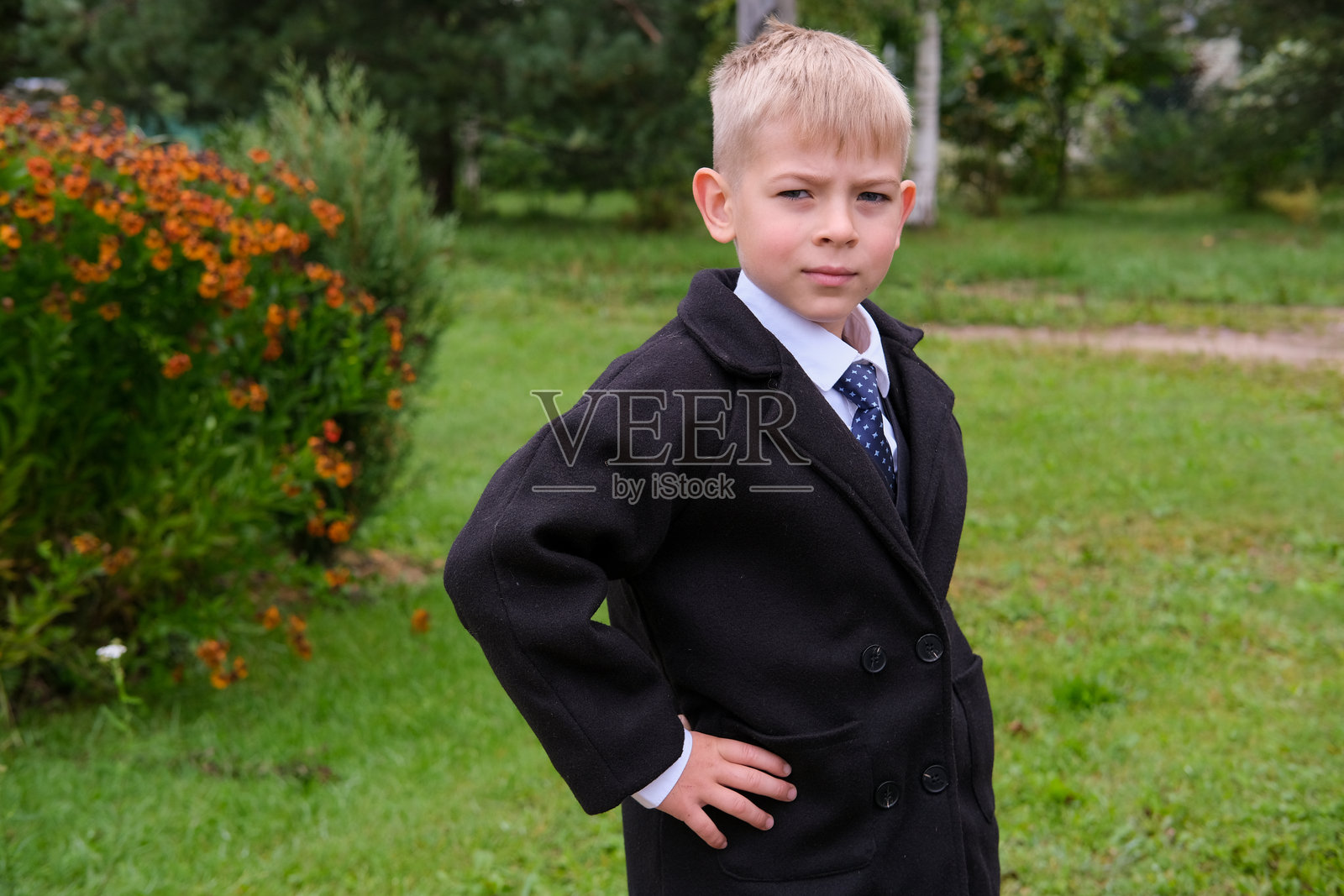 自信的男孩，身着时尚正装，在美丽的户外环境中摆姿势。男孩自信地穿着时尚的黑色西装，系着闪亮的领带，背景是郁郁葱葱、生机勃勃的绿色。照片摄影图片