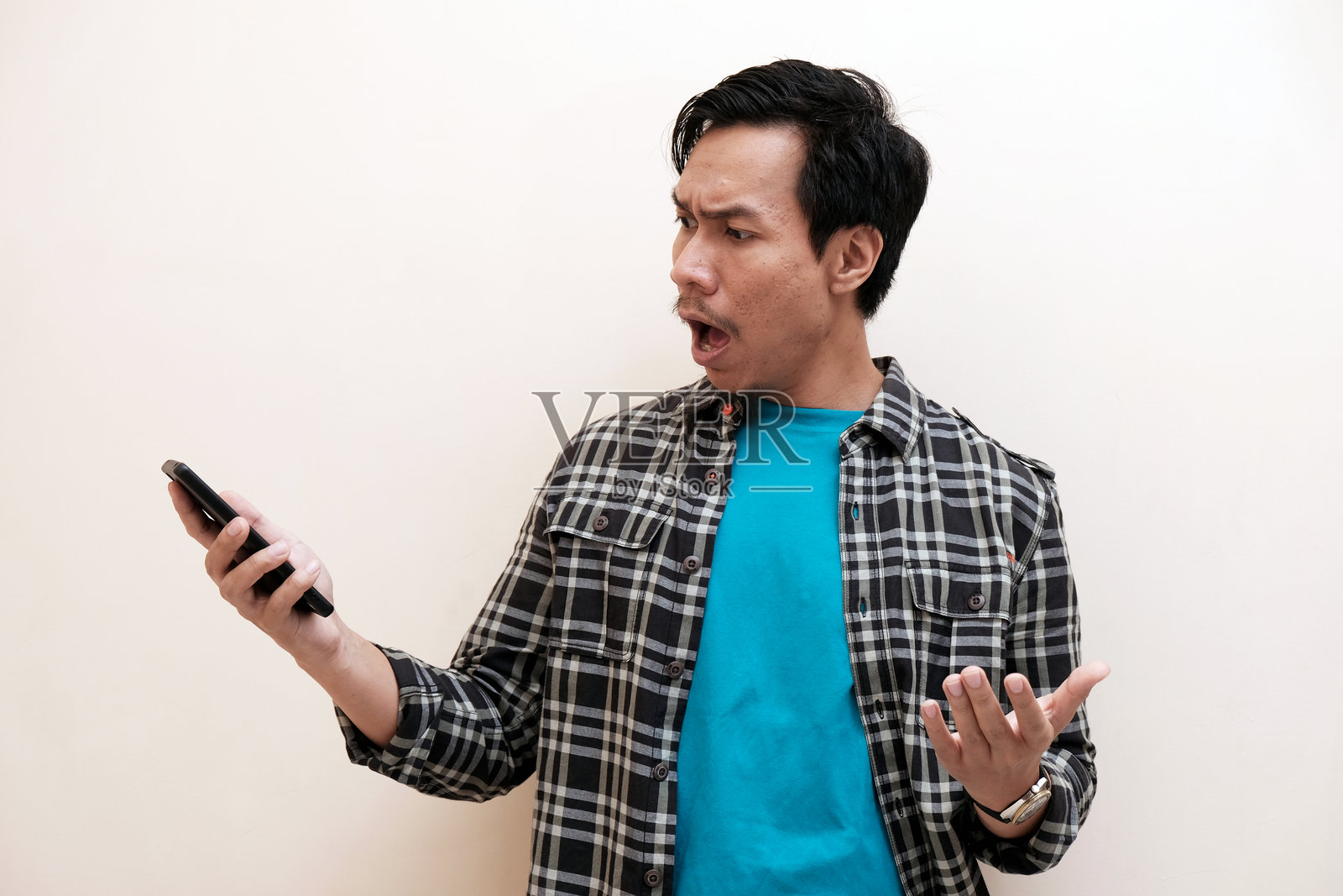 一位东南亚男子穿着法兰绒衬衫，手持手机，表情震惊地看着手机。照片摄影图片