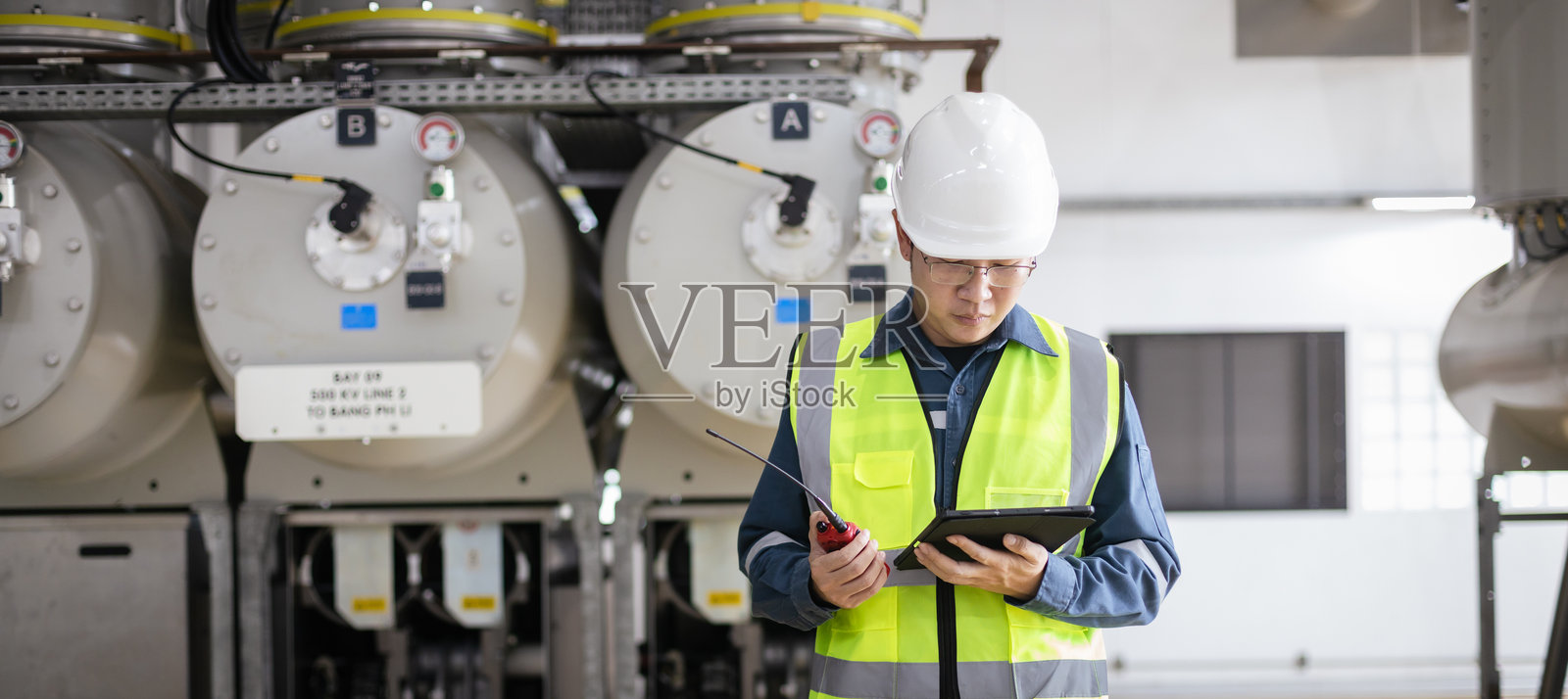 男性工程师正在检查高压气体绝缘开关设备，确保安全、控制和精确，他佩戴着安全帽和反光背心，并使用平板电脑和对讲机。照片摄影图片