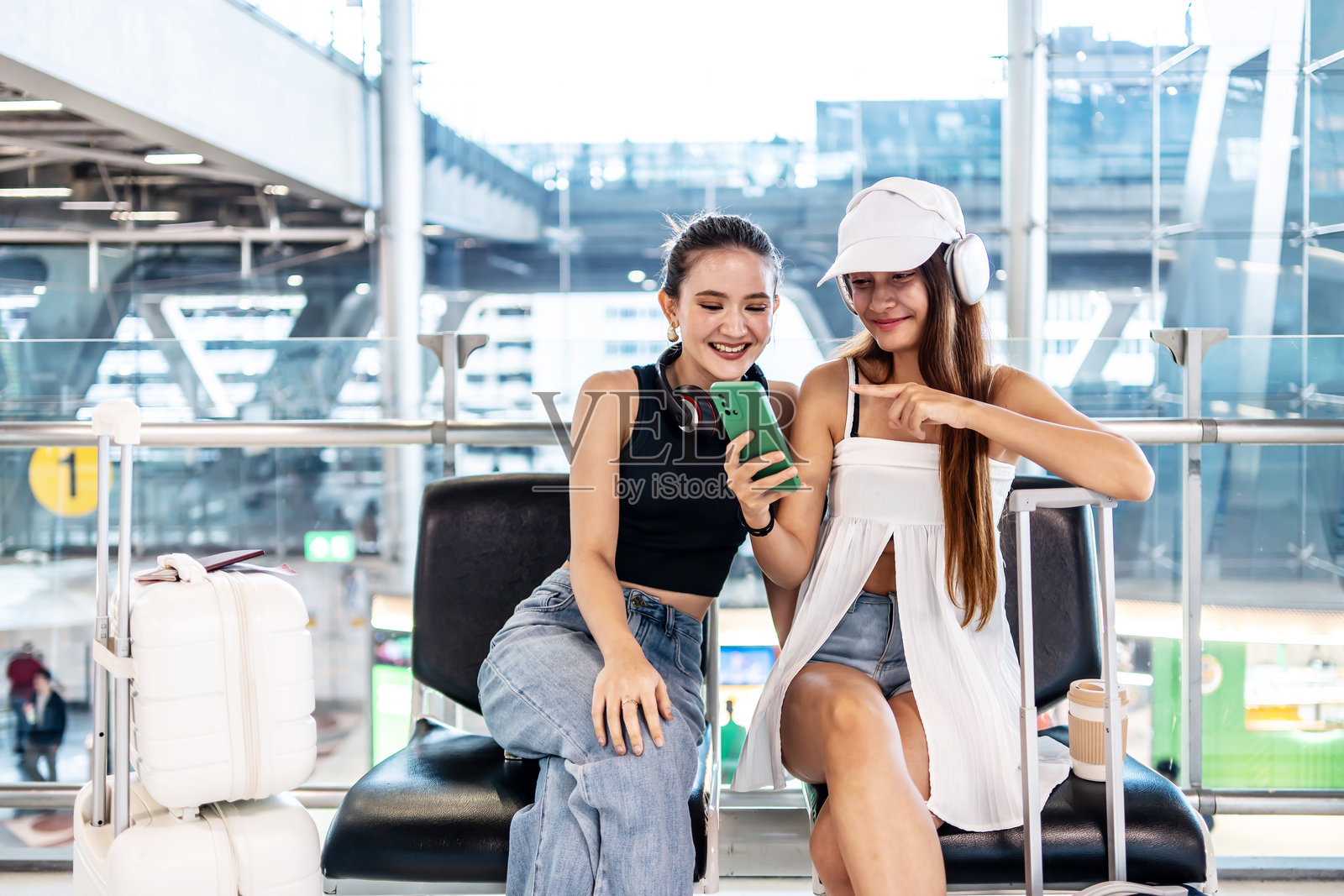 两位开朗的亚洲年轻女性朋友在充满活力的机场航站楼候机时，与她们最好的朋友进行有趣的视频通话聊天，边笑边聊。照片摄影图片