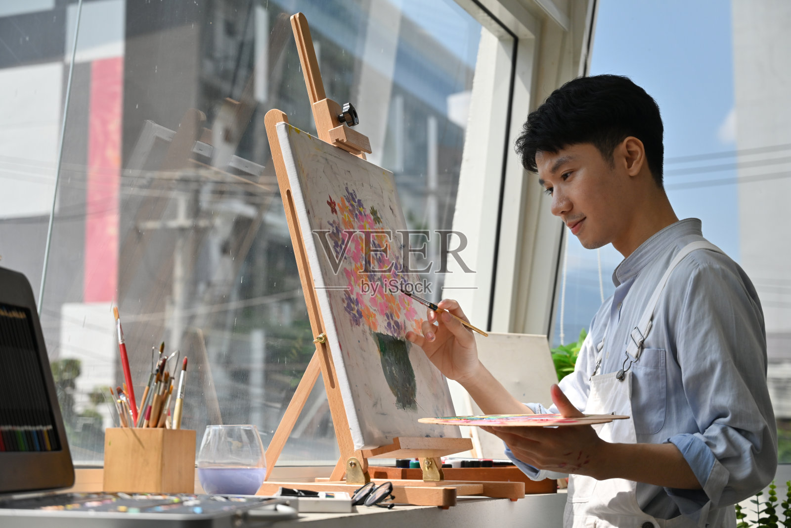 一位专注的年轻艺术家在明亮的画室空间里，临近一扇大窗户，在画布上绘制充满活力的花卉艺术作品。照片摄影图片