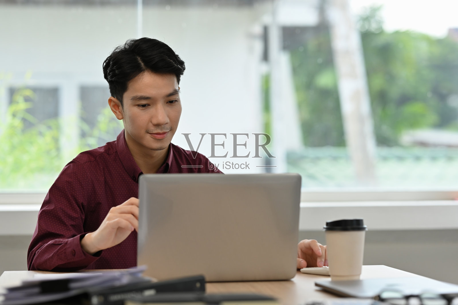 身穿酒红色衬衫的专注亚洲商人正在办公室里全神贯注地使用笔记本电脑，桌上放着咖啡。照片摄影图片