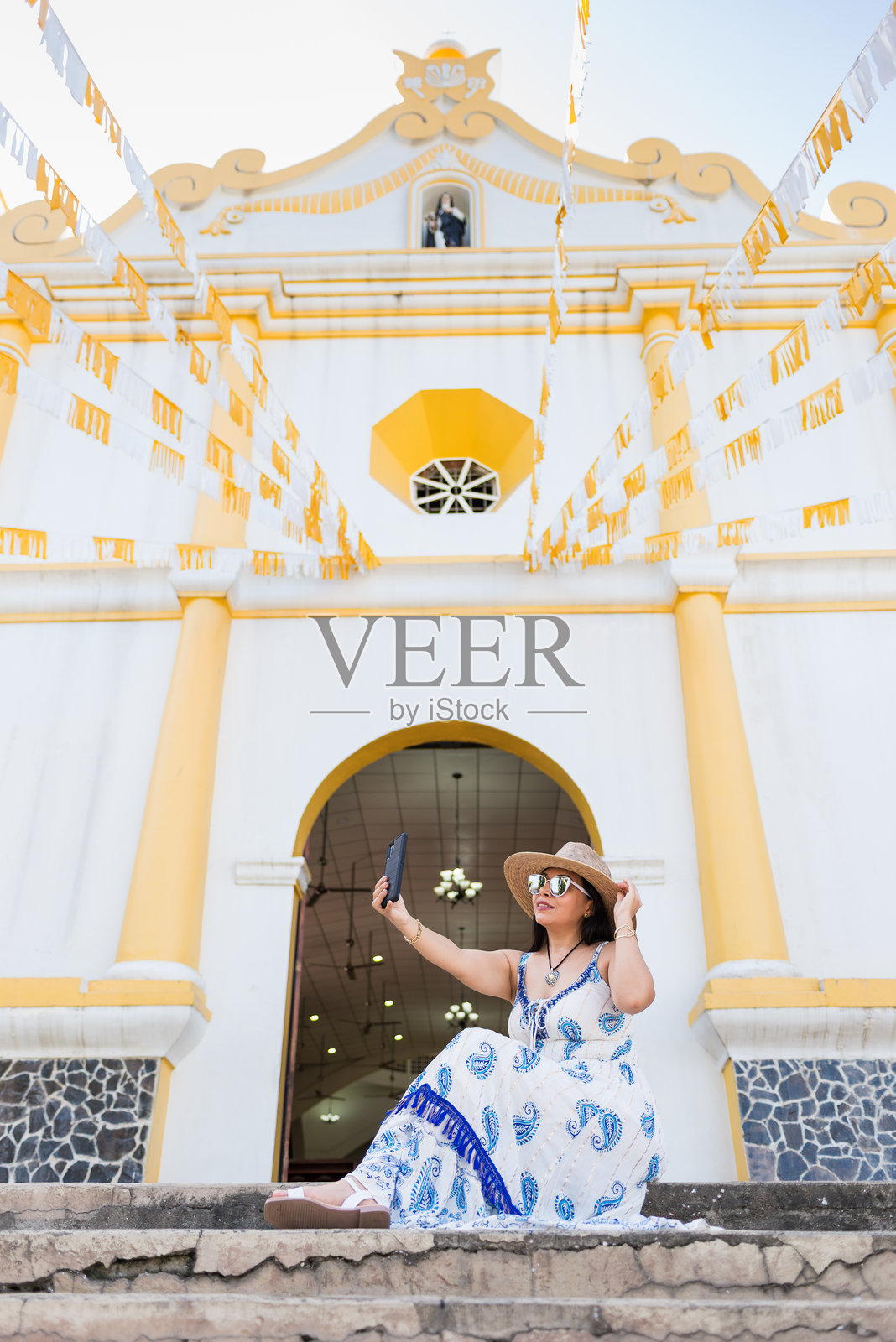 一位戴着帽子、穿着蓝白连衣裙的女士坐在教堂的台阶上自拍。照片摄影图片