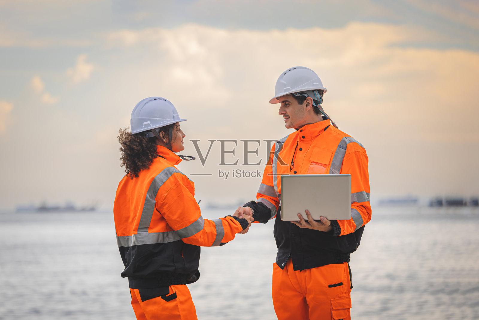 穿着安全帽和反光夹克的工业工程师在海滨港口握手，穿着安全装备和携带笔记本电脑的男性和女性工业工人在海边握手照片摄影图片