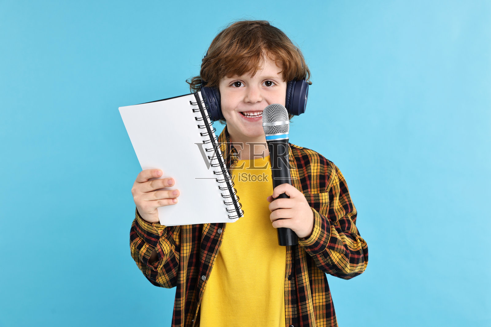 小男孩戴着麦克风、耳机，旁边放着笔记本，背景为浅蓝色。照片摄影图片