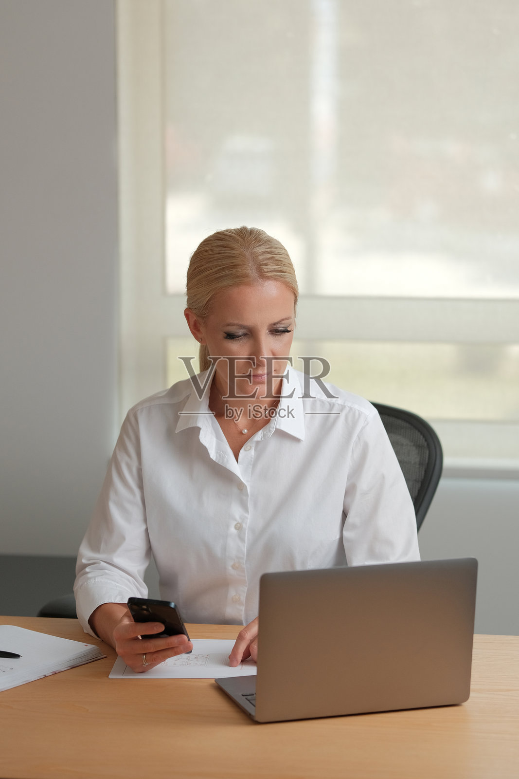 职业女性，身穿白衬衫，坐在办公桌前，在一本笔记本上写字，旁边放着笔记本电脑，办公室环境现代。照片摄影图片