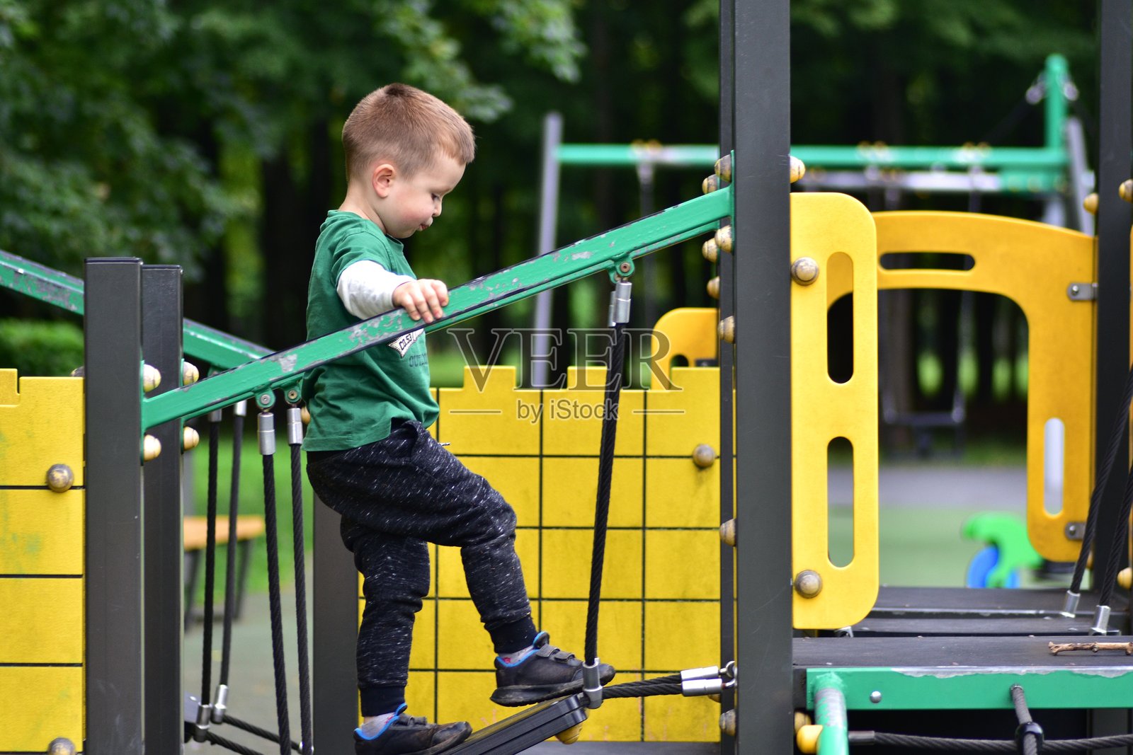 穿着绿色衬衫的男孩在游乐场的悬索桥上小心翼翼地行走。孩子在移动的桥上保持平衡，锻炼协调能力，享受户外乐趣。照片摄影图片
