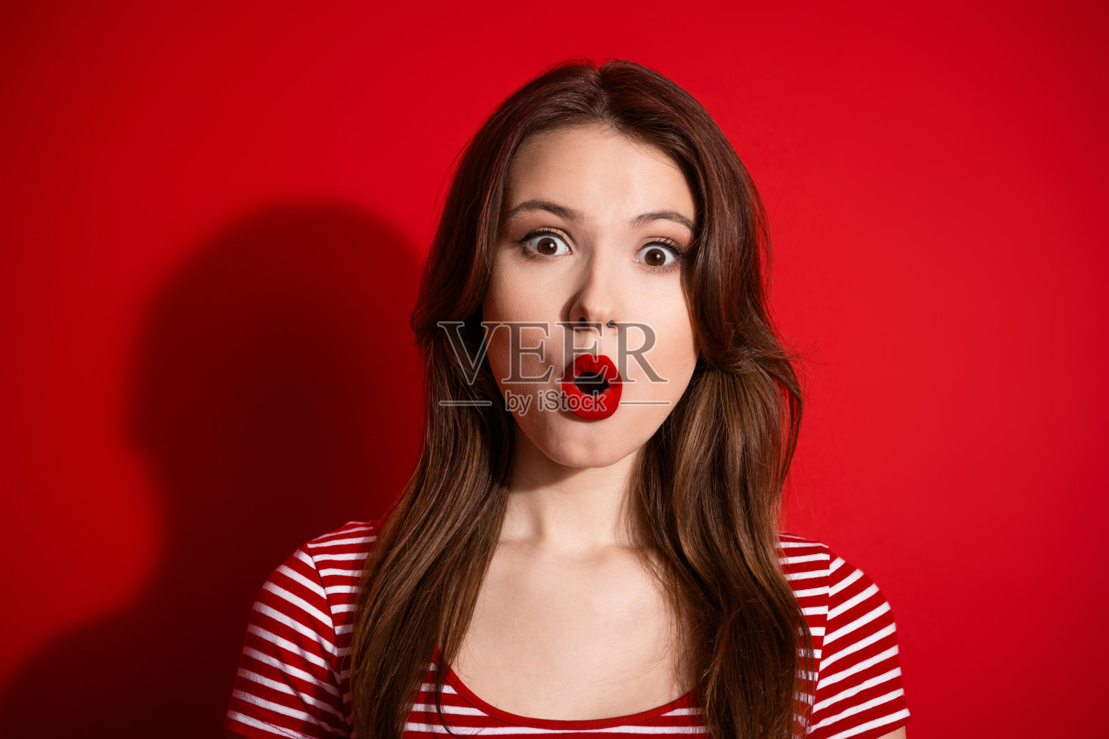 年轻女子惊讶地张着嘴，身穿休闲装，站在鲜红的背景前，她涂着鲜红的口红，表情丰富。照片摄影图片