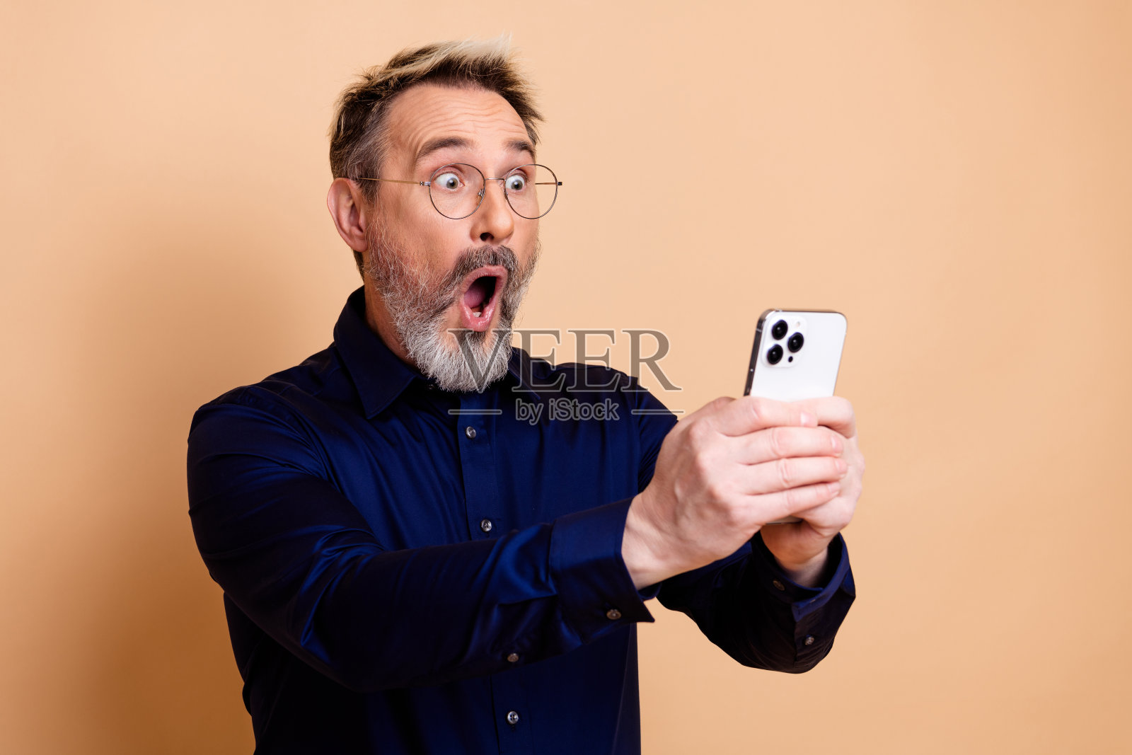 穿着正装的惊愕的中年男子，手持智能手机，背景为米色，脸上带着惊讶和好奇的表情。照片摄影图片
