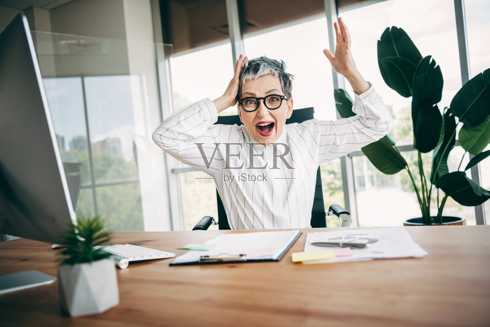 一位兴奋的成熟的女商人在现代办公室工作区庆祝成功，办公桌上有电脑和文件。照片摄影图片