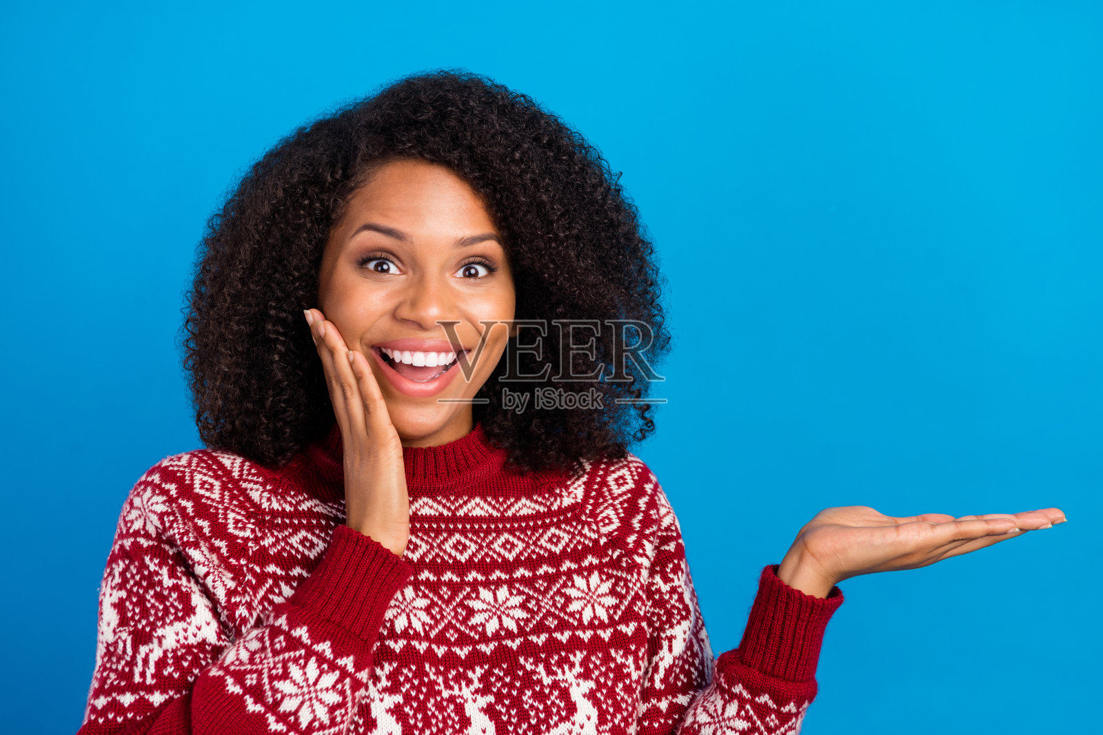 一位穿着印花毛衣的漂亮女子，一只手放在脸颊上，另一只手放在空出的空间，背景是孤立的蓝色。她看起来很惊讶。照片摄影图片