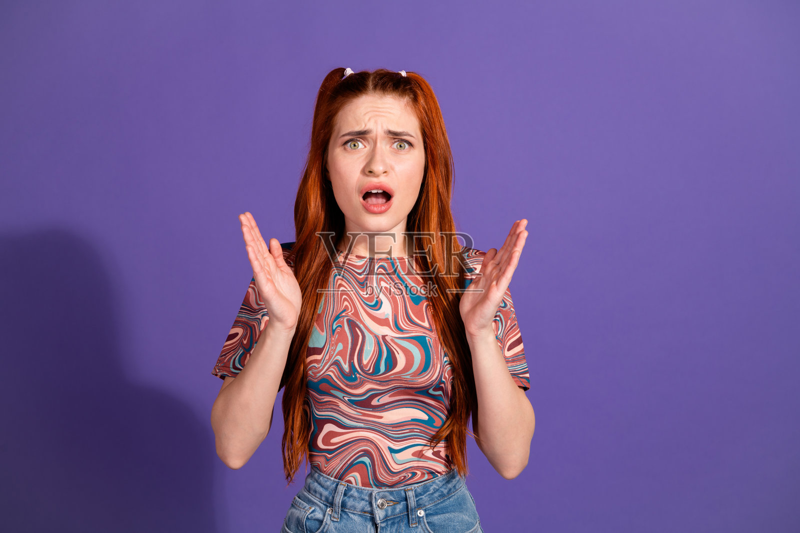 年轻女子，拥有鲜艳的红发，惊奇地看着镜头，背景为紫色，身穿时髦的抽象图案上衣照片摄影图片