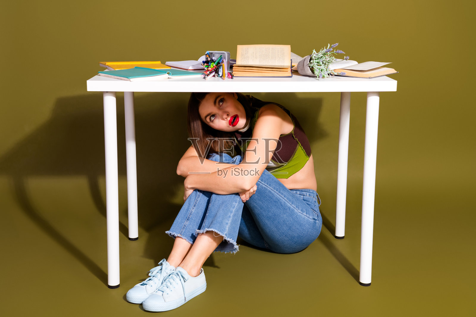 身着休闲装的年轻女子坐在书桌下，周围散落着书籍和文具，展现出时尚潮流的外观。照片摄影图片
