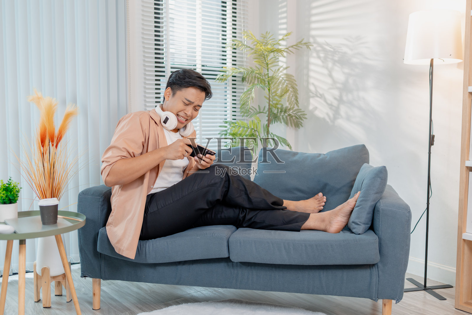 一个年轻男子在家里的沙发上坐着，穿着休闲服，脖子上戴着耳机，看着智能手机，脸上带着喜悦和兴奋的笑容，正在庆祝。照片摄影图片