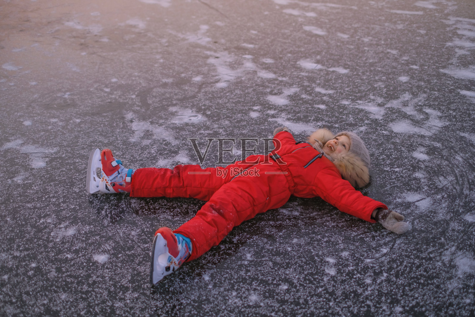 快乐大笑的女孩穿着红色连体滑冰服，脚穿冰鞋，躺在冰封湖面的冰上。户外冬季活动照片摄影图片