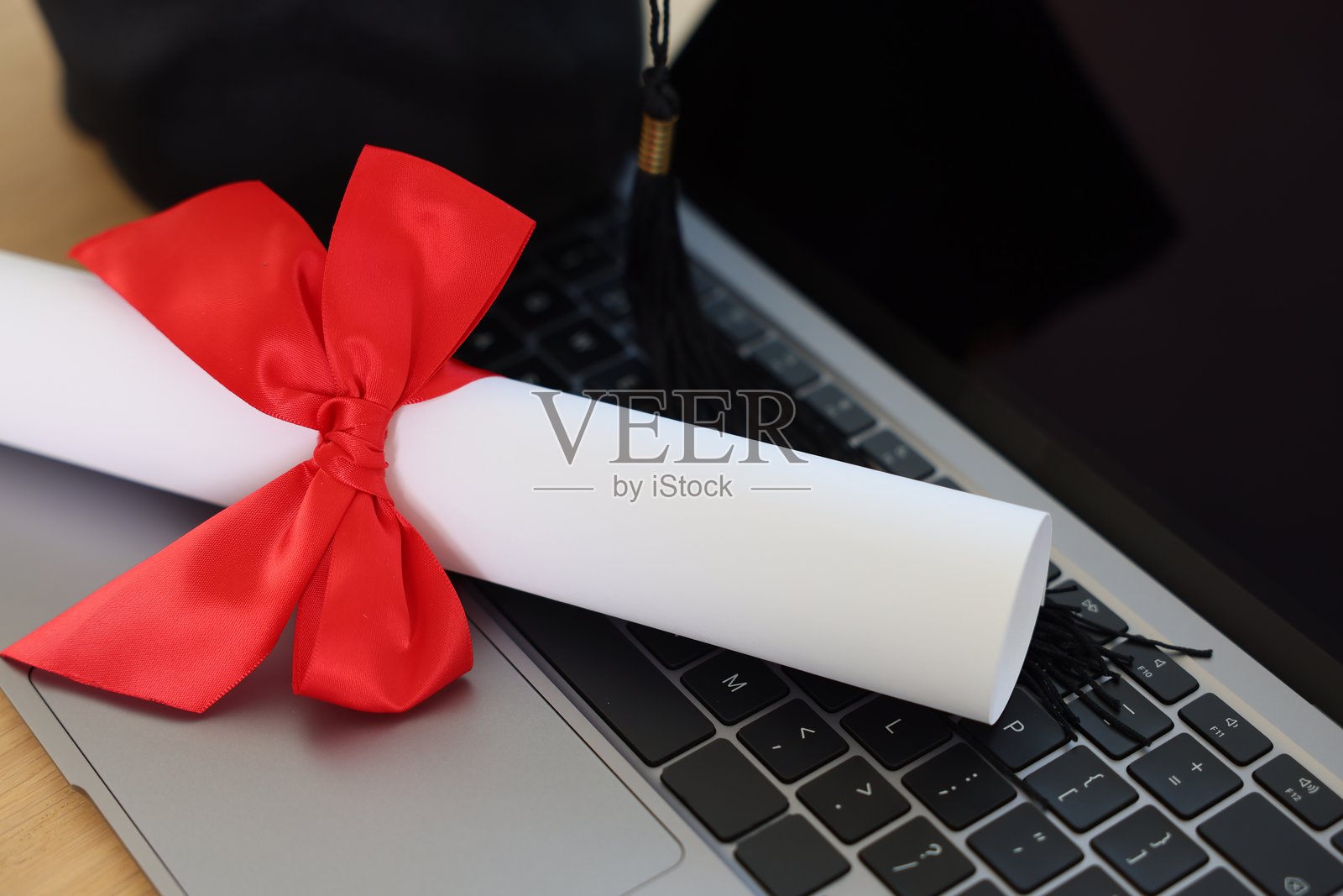 特写：打开的笔记本电脑和带有红色蝴蝶结的卷轴毕业证书放在木桌上照片摄影图片