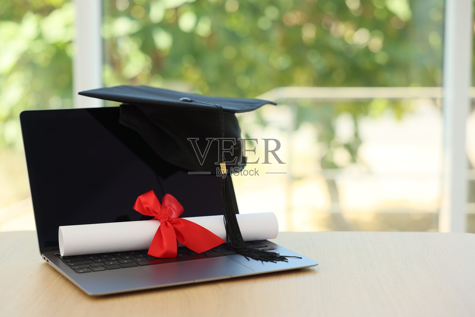 毕业帽、笔记本电脑和文凭放在木桌上，背景为模糊的绿色，特写。留有文字空间。照片摄影图片