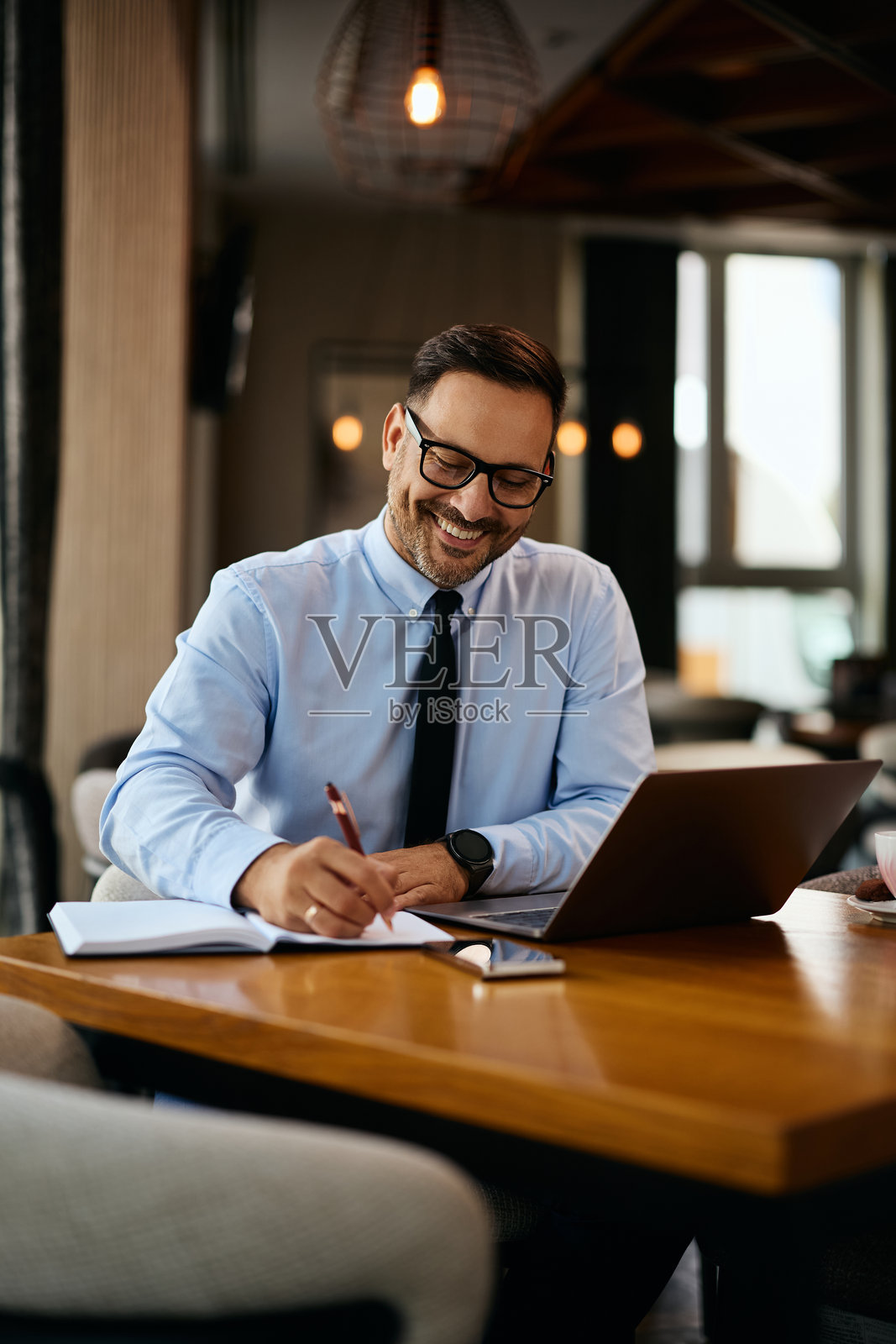 快乐的商人在咖啡馆用笔记本电脑工作时记笔记。照片摄影图片