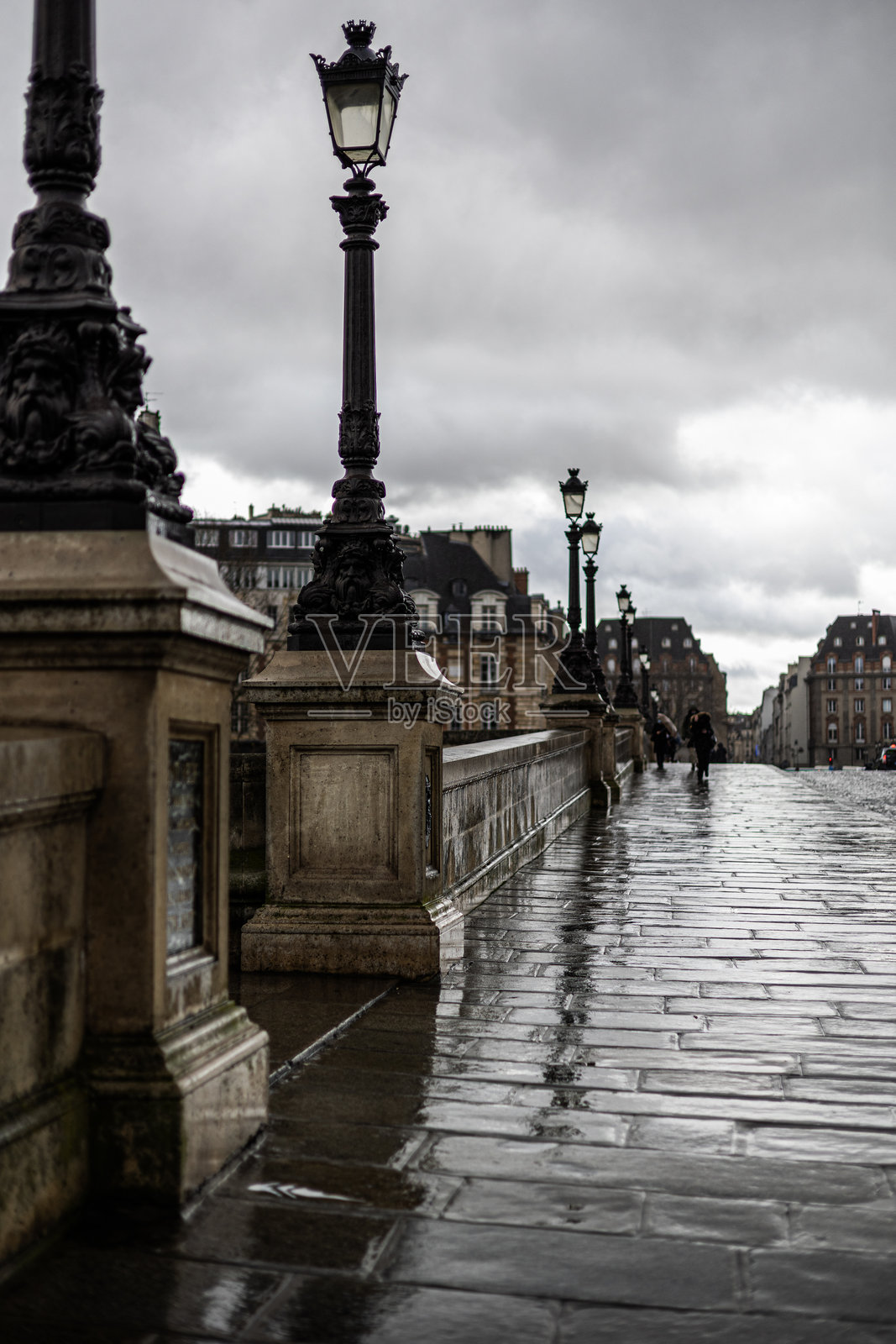 巴黎新桥上华丽的灯柱排列在潮湿、反光的石板路上。雨天阴沉，远处有人在行走。照片摄影图片
