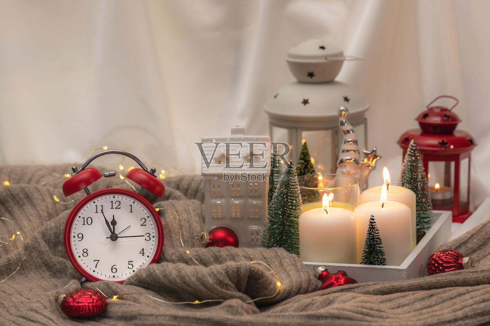 木质托盘上燃烧的蜡烛和圣诞灯笼，配有温暖的针织毯子和红色圣诞球。红色闹钟，差五分十二点。圣诞、新年背景。焦点区域。照片摄影图片