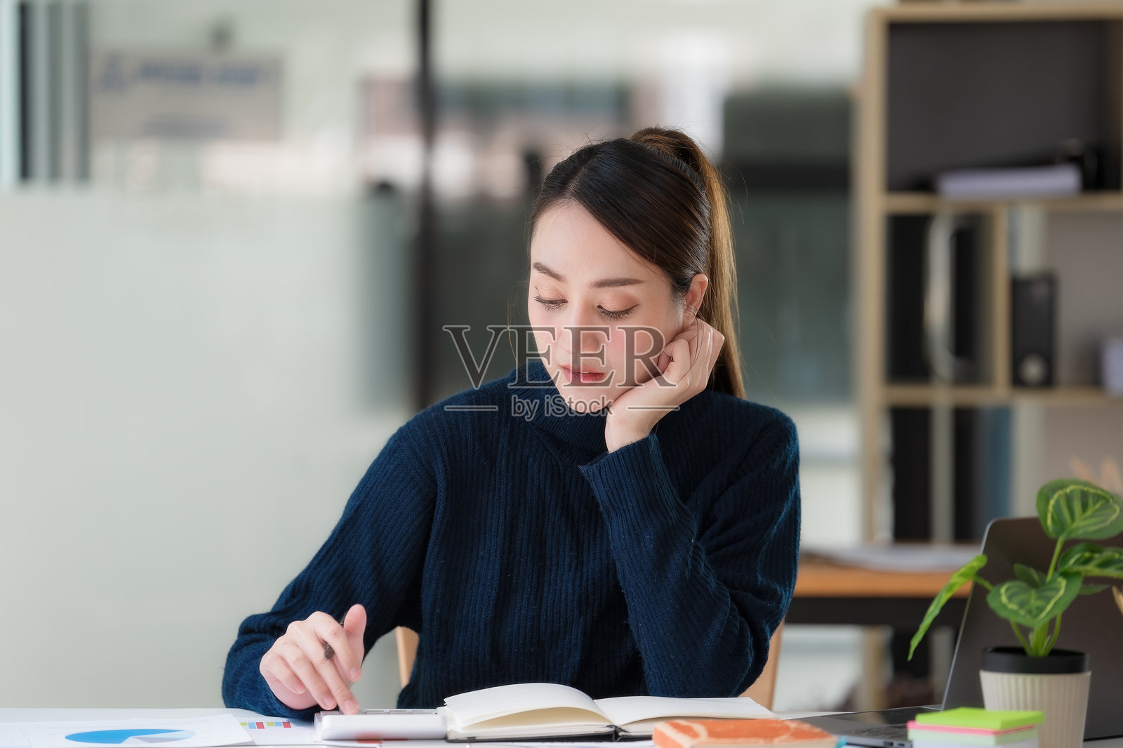 一位年轻的亚洲女商人坐在现代办公室的办公桌前，在笔记本上思考并书写。照片摄影图片