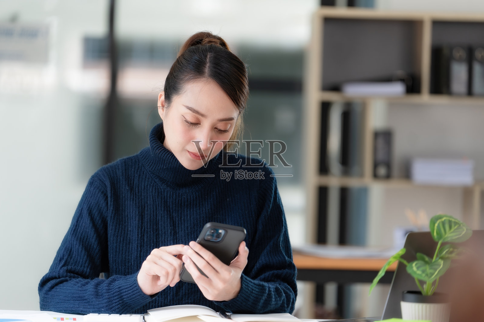 亚洲女性坐在办公室的桌子旁，一手拿着手机，一手在打字。照片摄影图片